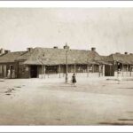 Image: Three girls in late 19th century dresses play in a wide dirt road while older women stand together outside stores with wooden shingled roofs and verandahs.  In the centre of the image a gas street light can be seen.