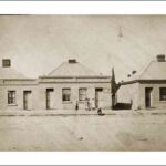 Image: a group of three pairs of semi-detached simple fronted cottages with a single door and window each and a central chimney rising from each roof. In front of the buildings stand women and children in late 19th century clothing.