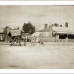 Image: a group of men, women and children in late 19th century clothing gather around a horse drawn cart on a dirt road lined by single storey cottages.