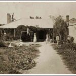 Image: A family (a man, woman, three boys and two girls) and their dog pose in an overgrown garden outside a single storey cottage with verandah.