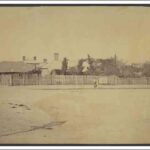 Image: fruit trees, tin roofs and brick chimneys rise above a paling fence bordering a dirt road. A number of people in late 19th century dress can be seen standing in front of the fence.