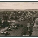 Image: A wide city street is lined with large buildings and a power station on one side and parklands on the other. A number of horse drawn vehicles are using the street.