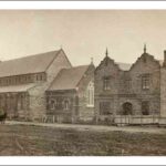 Image: a stone church with a clerestory roof sits along side a two storey rectangular stone building with two gable roofs with scalloped ends topped with finials on the street side.