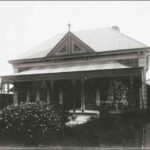 Image: A symmetrical fronted bluestone villa with decorative wrought iron verandah, tin roof and decorative gable end with finial centred above the front door sits in a established garden with a number of flowering bushes.