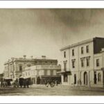 Image: horse drawn cabs for hire stand in the centre of a dirt city road which is lined with two and three storey public buildings