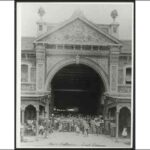 Image: A large group of people men, women and children pose under a huge two-storey high archway with a decorative pediment set within a brick terrace building fronted on either side of the arch with verandahs.