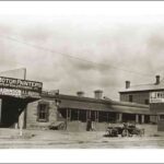 Image: a street-scape with a single storey stone workshop on the left, a hand barrow in front of it, a terraced row of cottages with verandahs in the centre of the image, a 1920s truck parked in front, and a two storey building to the right.