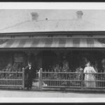 Image: a man and a woman stand behind a wooden fence in front of a bluestone cottage with tin roof and verandah. The man stands at the gate and wears a dark suit and bowler hat, the woman who is to the right of the photograph wears a white dress.