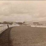 Image: A man leans against a high picket fence lining a curved racecourse. Two  grandstands can be seen in the distance.