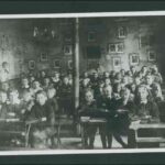 Image: a large group of boys and girls in early 20th century clothing pose sitting at their desks in a school room. In the background their teachers watch over them.