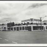 Image: a single storey concrete building with a flat roof on the corner of two main roads. The building is being used as a health studio with signs advertising a pool inside. 1970s era cars are parked outside.