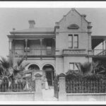 Image: A woman in a white dress and her small dog stand in front of a two storey house with a bay window, arched verandah and balconies.