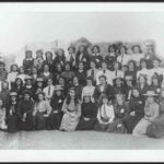 Image: a large group of girls in early 20th century dresses, some wearing straw hats, pose in four rows for this formal school photograph.