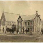 Image: a gothic style church with arched porch and clerestory windows and associated two-storey manse made of the same dark stone with arched windows and portico.