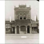 Image: A single fronted two storey building dominates this view. It is built from rendered stone with cement dressings and arched doorway and windows. It features a stepped parapet with decorative urns and the inscription "ESTb 1893".