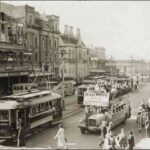 Image: a city street busy with pedestrians, electric trams and buses. In the left foreground is the Adelaide-Glenelg Railways Bus, a double decker with an open top, its passengers enjoying the sunshine. One of the buildings is in process of remodelling.