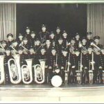 Image: A large, all-male brass band poses with their instruments on a stage