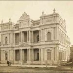 Image: a highly decorative three storey stone public building in a French Renaissance style with a front entrance flanked by columns supporting a triangular pediment, balconies, and parapets with stone urns and a carved lion and unicorn coat of arms.