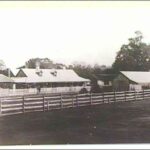 Image: Black and white photograph of homestead with outbuildings and a wooden fence in foreground