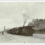 Image: passengers board a steam train which is stopped in the middle of a city street