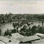 Image: A floating building with a multi-domed roof sits on a lake surrounded by trees and lawns. A rotunda can be seen on the shore by the floating building and in the background, across the lake, churches and houses can be seen.