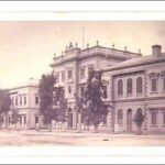 Image: a large public building. The centre is three-storeys high with a flagpole on the flat roof and stone urns decorating the parapet. The wings on either side are two storeys high and have low pitched roofs.