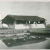 Image: A wooden framed building with a thatched roof and half height mud-brick walls stands behind a square edged pool in a barren field