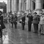 Image: a small group of men in military dress uniforms stand saluting on the pavement in front of a set of stone steps leading up to a building with columns.