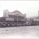 Image: a row of 1920s era cars are parked on a 45 degree angle facing a dirt road. Behind them is a single storey commercial building, part of a row of terraced shops, with a curved parapet and a sign which reads "Autocars Limited".