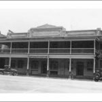 Image: A two storey hotel with balcony set within a terrace. 1920s era cars are parked on the street outside.