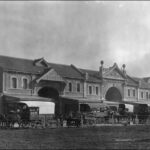 Image: A long two-storey brick terrace building with three large double height arches and decorative gables. Along the street at the front a number of horse drawn carts are parked.