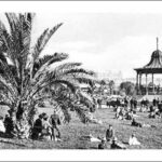 Image: A crowd of people in early twentieth century attire sit on the grass around a rotunda where a band plays