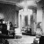 Image: late 19th century drawing room with high ceilings, a large chandeleir and heavily-patterned wallpaper and furniture