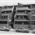 Image: Three 1980s era cars are parked in front of four-storey 1980s "modern" brick townhouses with angular glass railed balconies.