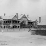 Image: A very large house on a corner block set in an established garden behind a stone wall. The house, with arched verandahs, balconies and at least five chimneys is half covered in ivy.