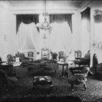 Image: A stately drawing room with luxurious carpets and curtains, a large fireplace, and a rang of chairs and tables of different shapes and sizes.