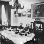 Image: The dining room of a large house in the 1890s. The room has a number of paintings on the wall, a bay window, and the table is set for eight.