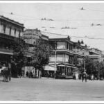 Image: a series of three storey buildings line one side of a wide city street. Between the buildings and the road is a line of trees. Travelling along the street are horse-drawn vehicles, motor cars and an electric tram, as well as people in 1920s clothes