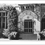 Image: Front entrance to large stone house. Stone steps lead up to the arched front door, flanked by two columns. To the left a window is completely surrounded by ivy and to the right is a verandah with decorative wooden fretwork.