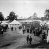 Image: a crowd of men and women in early 1870s clothing stand amongst tents and agricultural displays. In the background a large building can be seen.