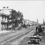 Image: Horses and buggies travel down a wide dirt road. In the distance two towers (the Town Hall and Post Office) can be seen on either side of the street.