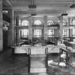 Image: the interior of a bank which is lined with marble and features a mezzanine balcony level, double height arched windows and square columns