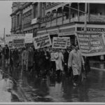 Image: men in 1940s era clothing march through the rain holding protest signs demanding increased rights for workers