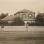 Image: a two storey stone house behind a high fence and partly obscured by trees. The second story windows are shuttered and an arched verandah supports a balcony with stone balustrade.