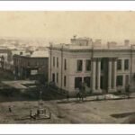 Image: a two storey stone building with a rectangular floorplan and an entrance flanked by huge, two storey columns stands on a street corner and surrounded by other commercial and public building