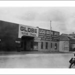 Image: a man in a 1930s era coat poses on a motorcycle outside a shop with a rectangular arched entrance and sign reading "Globe Motor Park"