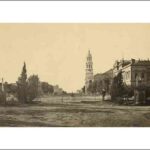 Image: a black and white photograph of a view across a city park and down a wide dirt street. Lining the street are a number of large public buildings including one with a large tower.