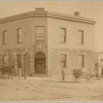 Image: a two storey corner hotel with an exposed stone façade on its lower floor and a rendered upper floor. A lantern hangs over the arched door on the corner, a double-hung window above with two more windows per floor on each side of the building.