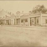 Image: a row of single and double storey terraced buildings with a variety of rooflines, including arched parapets and exposed gable ends, fronting a dirt road.