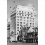 Image: a large building of at least ten storeys with a square floor plan, flags flying from poles on the roof, and arches on the ground floor.